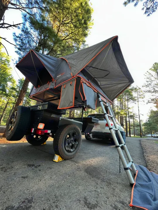 This rv rental setup features a deployed rooftop tent on an off-road camper trailer in a wooded campground.. Ady ORT 2022