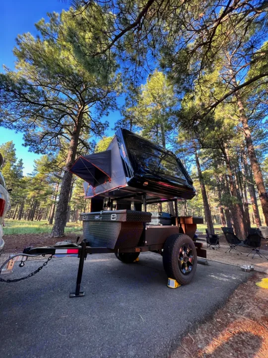 A dark overland trailer with a deployed rooftop tent, an RV rental alternative, parked at a forest campsite.. Ady ORT 2022