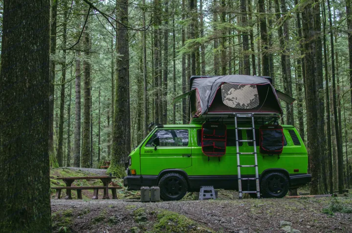 Explore nature with this green camper van featuring a rooftop tent, parked in a serene forest campground. Great RV rental idea!. Volkswagen Vanagon 1989