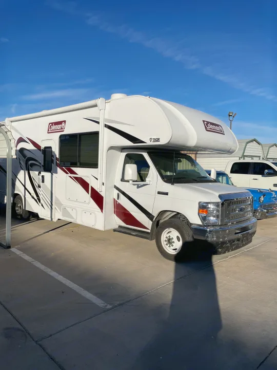 White Coleman Class C motorhome for RV rental, parked on a sunny day. Features red/black decals and a large shadow in front.. Thor Coleman 2022