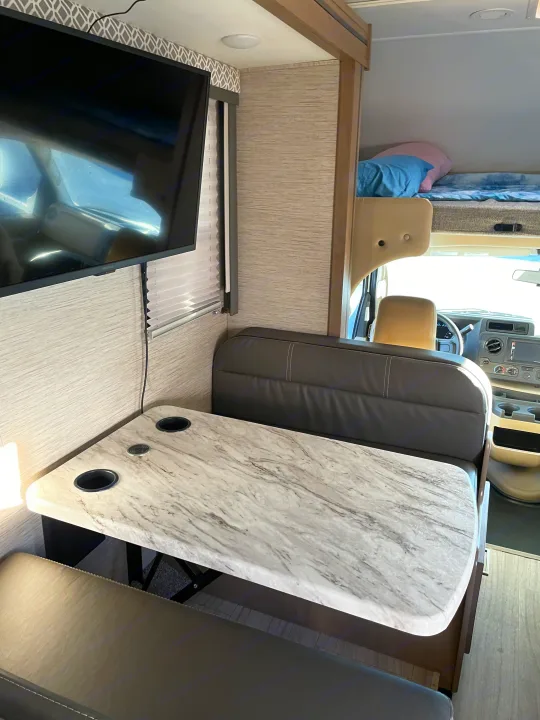 Modern RV rental interior with dinette, marble-look table, TV, and an overhead bunk bed above the cab area.. Thor Coleman 2022