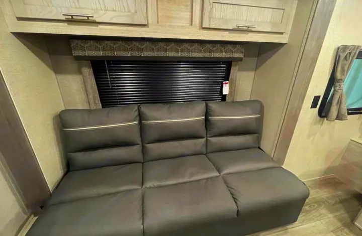 Comfortable gray RV rental interior featuring a three-seat sofa, overhead cabinets, and a window with blinds.. Coachmen Catalina 2022