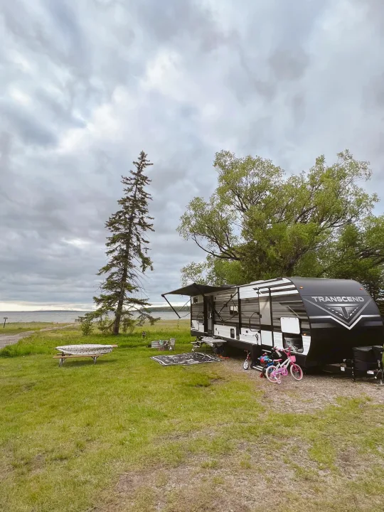 This RV rental, a Transcend travel trailer, is set up at a spacious lakeside campsite with children's bikes, under a cloudy sky.. Grand Design Transcend Xplor 2022