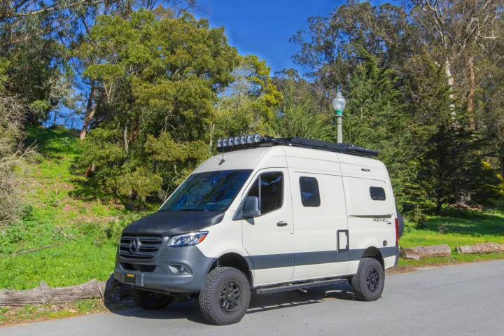 White Mercedes-Benz Revel 4x4 camper van, ideal for an rv rental, parked on a road with green hills and trees.. Other Revel 2023