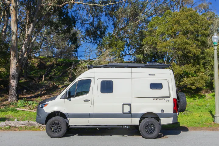 A cream Sprinter camper van, perfect for RV rental adventures, is parked outdoors. It features off-road tires, a roof rack, and a rear spare.. Other Revel 2023