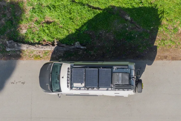 Top-down view of an RV rental van featuring rooftop solar panels and adventure gear, parked on asphalt next to a grassy area.. Other Revel 2023