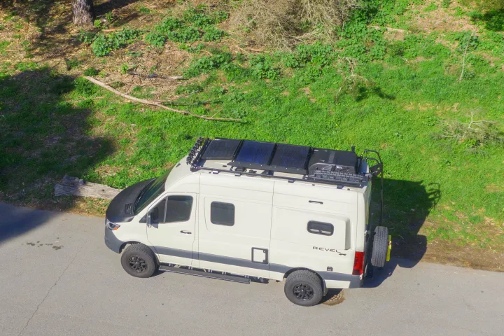 White off-road Sprinter camper van, equipped with solar panels and roof rack, parked by a hill. Great rv rental option.. Other Revel 2023