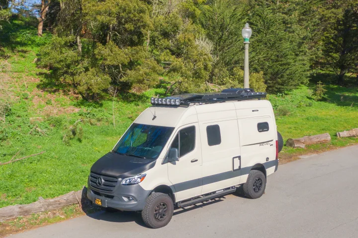 This adventure-ready Mercedes Sprinter camper van, perfect for rv rental, is parked on an asphalt road by a lush, treed hillside.. Other Revel 2023