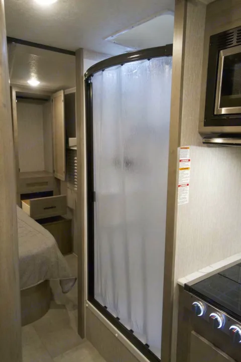 Compact RV rental interior: kitchen with microwave/stovetop, modern curved shower, and bedroom storage area.. Coachmen Freelander 2023