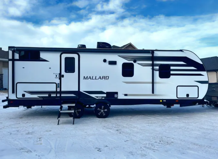 A white Mallard travel trailer, often offered as an RV rental, sits on snow under a blue sky.. Heartland RVs Mallard 2021