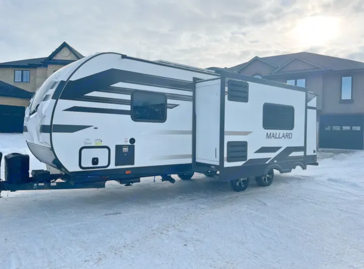 A white and black Mallard travel trailer, ideal for an RV rental, is parked on a snowy residential street.. Heartland RVs Mallard 2021