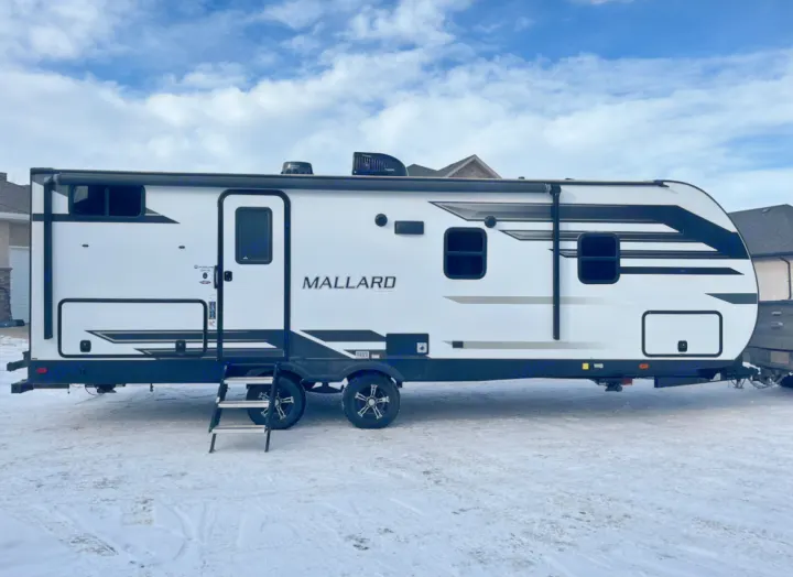 A clean white Mallard travel trailer with black accents is parked on snowy ground under a blue sky, ideal for an RV rental.. Heartland RVs Mallard 2021