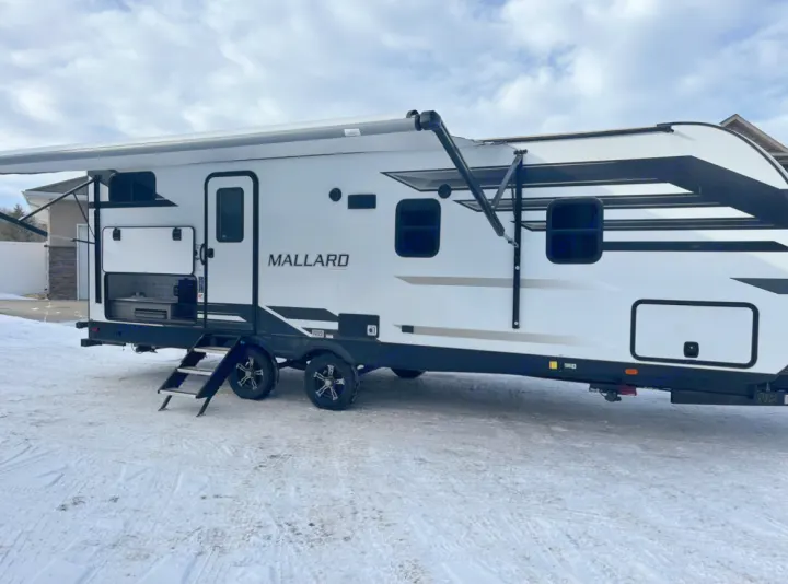 White Mallard travel trailer, an ideal RV rental, parked in snow with its awning extended and outdoor kitchen open.. Heartland RVs Mallard 2021