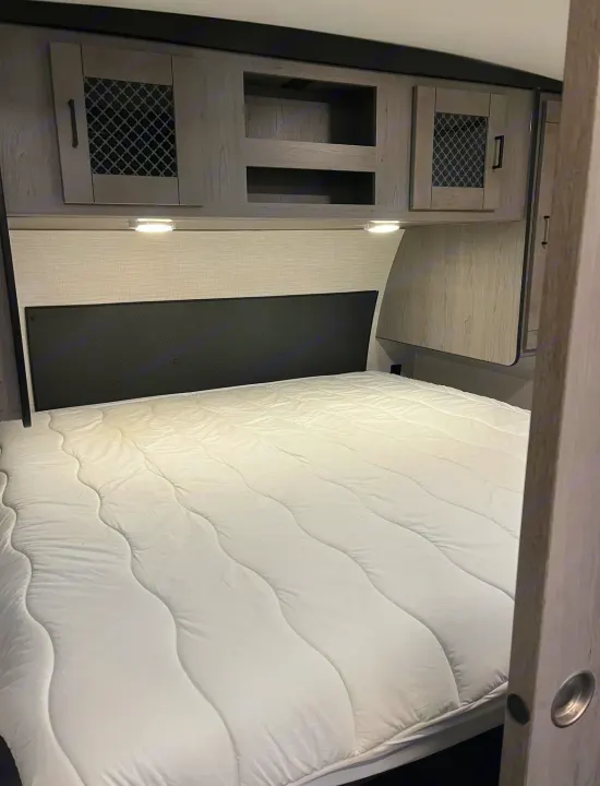 Inside an RV rental: a comfy bed with black headboard, overhead storage, and reading lights in a modern camper bedroom.. Heartland RVs Mallard 2021
