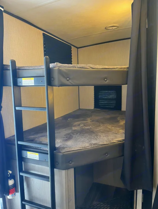 Inside an RV rental, showing compact bunk beds with a ladder & soft grey blankets, ideal for family sleeping quarters.. Heartland RVs Mallard 2021