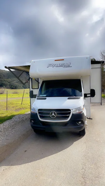 White Mercedes-Benz Coachmen Prism RV rental on a road, awning out, against a backdrop of green hills.. Coachmen Prism 2023