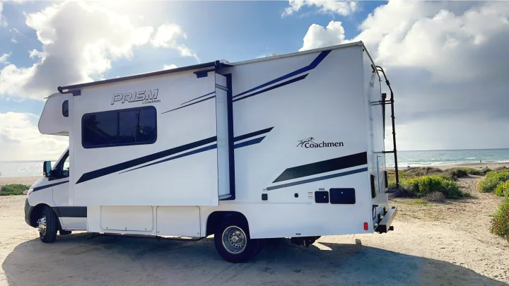 White Coachmen Prism RV rental parked on a sandy beach near the ocean under a cloudy sky, ideal for coastal travel.. Coachmen Prism 2023
