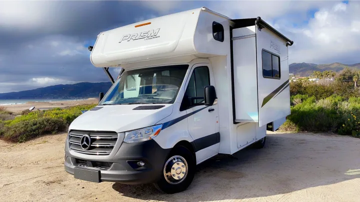 White Mercedes-Benz Sprinter RV rental parked on a sandy area by a beach, with mountains and cloudy sky.. Coachmen Prism 2023