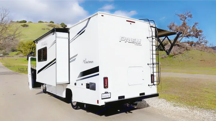 White Coachmen Prism RV rental parked on a road beside green hills. Slide-out extended, awning out.. Coachmen Prism 2023
