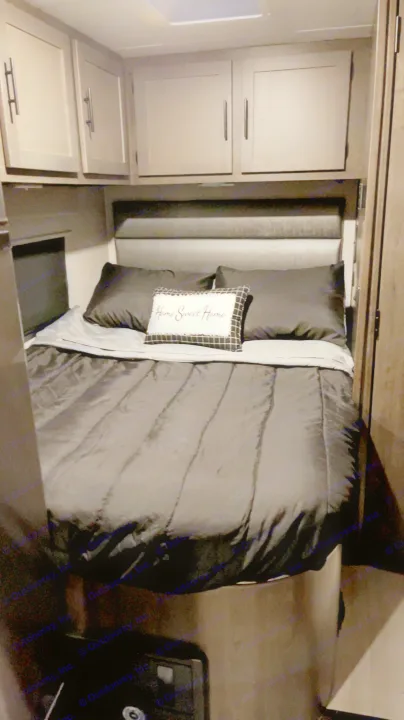 Inside this RV rental bedroom: a cozy space with a bed, dark bedding, "Home Sweet Home" pillow, and overhead cabinets.. Coachmen Prism 2023