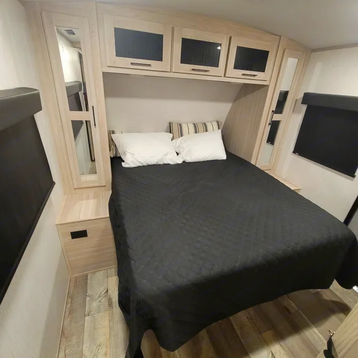 Comfortable RV rental bedroom with a dark comforter, white and striped pillows, and light wood cabinetry with dark glass inserts.. Heartland RVs Sundance Ultra Lite 2022