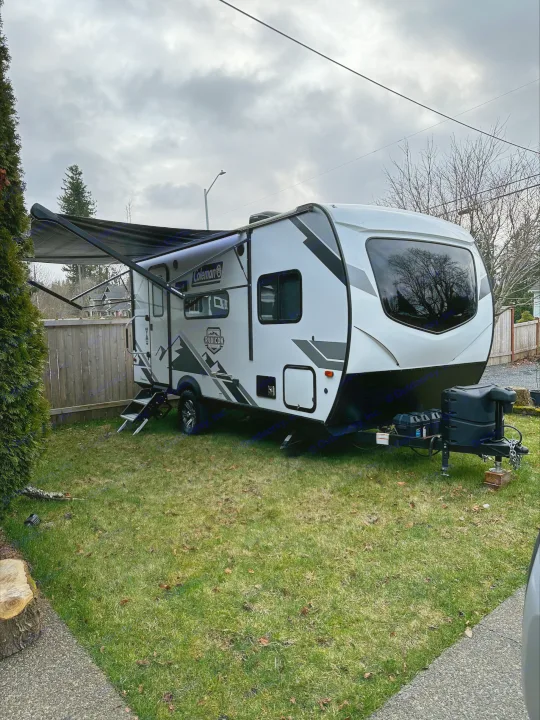 A modern white & black Coleman travel trailer, ideal for RV rental, parked on grassy lawn with lighted awning extended.. Dutchmen Coleman Rubicon 2021