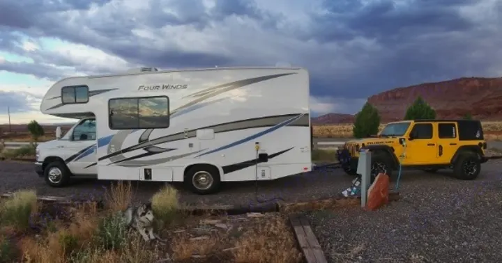 RV rental setup: white motorhome, yellow Jeep, husky dog at desert RV park with hookups under cloudy sky.. Thor Four Winds 2022