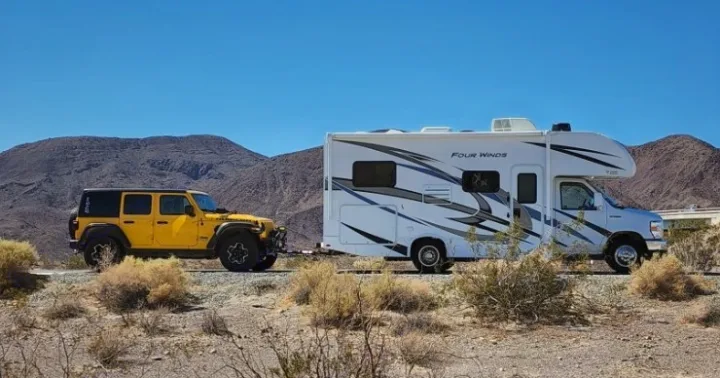A large white RV rental flat-tows a yellow Jeep Wrangler on a clear day through the desert mountains.. Thor Four Winds 2022