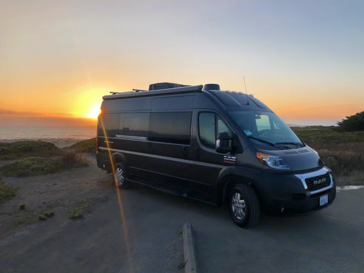 Golden hour RV rental adventure: dark gray camper van overlooking a vibrant ocean sunset by coastal land.. Thor Sequence 2021