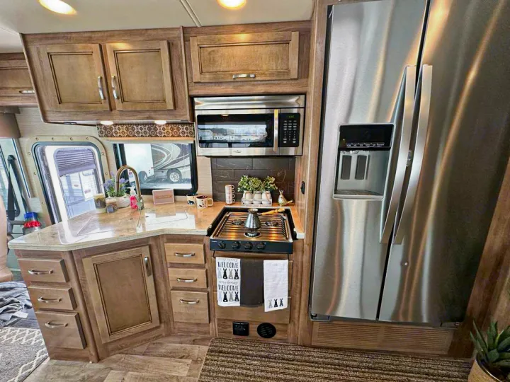 Modern RV rental kitchen featuring a large fridge, microwave, gas stove, and sink with wooden cabinets. Ready for travel!. Jayco Precept 2019