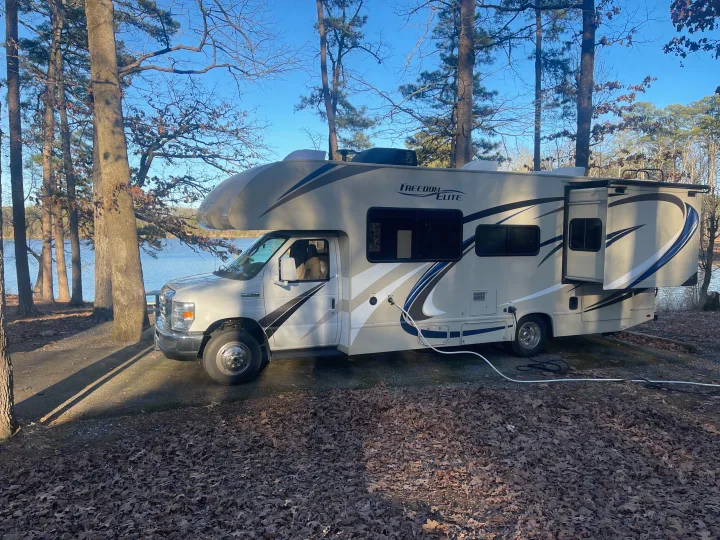 An RV rental (Freedom Elite motorhome) parked lakeside among trees under clear blue skies, ideal for a camping getaway.. Thor Freedom Elite 2019