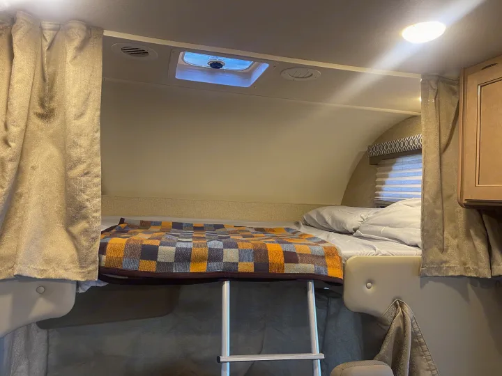 Cozy overhead bed in an RV rental, complete with a ladder, checkered blanket, and ceiling vent for comfortable mobile living.. Thor Freedom Elite 2019