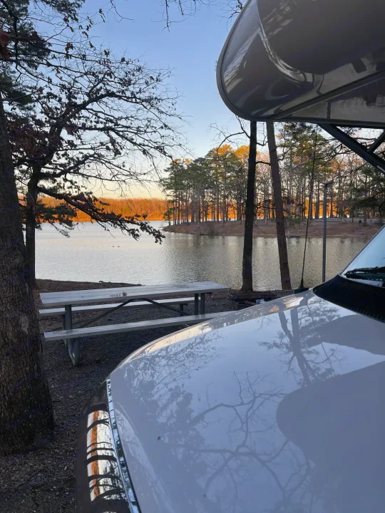 View from an RV rental at a serene lakeside campsite, with a picnic table and trees reflecting on the vehicle's hood.. Thor Freedom Elite 2019