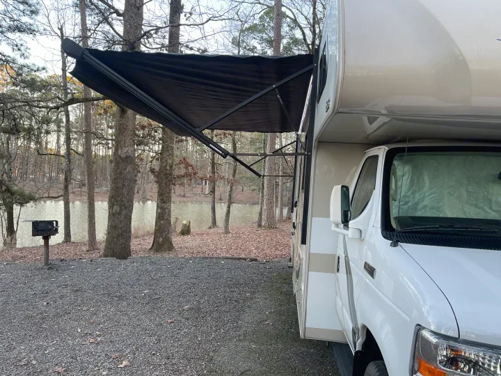 A white RV rental with its awning extended, parked on a gravel campsite next to a tranquil lake surrounded by trees.. Thor Freedom Elite 2019