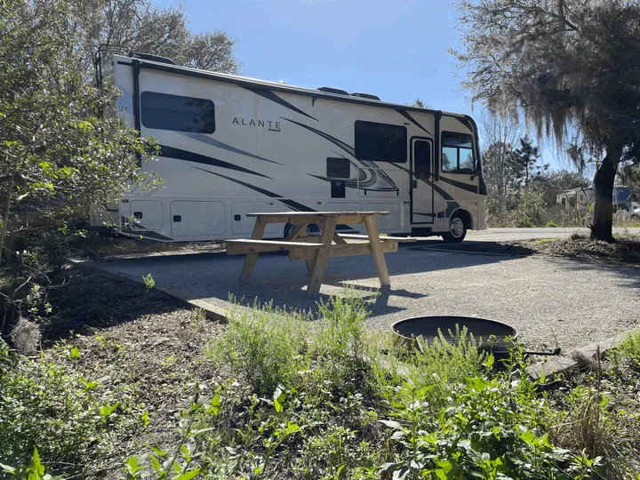 A white Alante Jayco RV rental rests at a sunny gravel campsite with a wooden picnic table and a black fire pit.. Jayco Alante 29F 2023