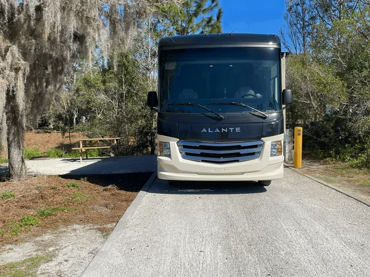 Front view of a two-tone Alante RV rental at a sunny campsite with a picnic table and fire pit nearby.. Jayco Alante 29F 2023