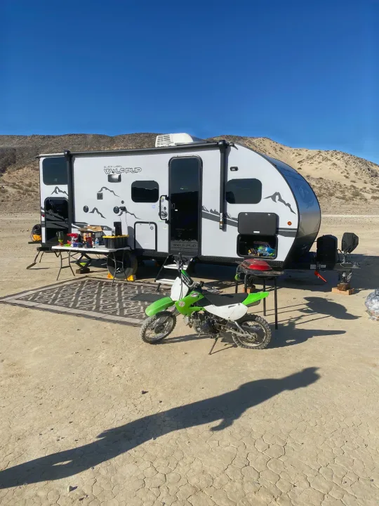 White RV rental in a desert camp with a green dirt bike, outdoor rug, and table setup. Clear blue sky and mountains.. Forest River Cherokee Wolf Pup 2022