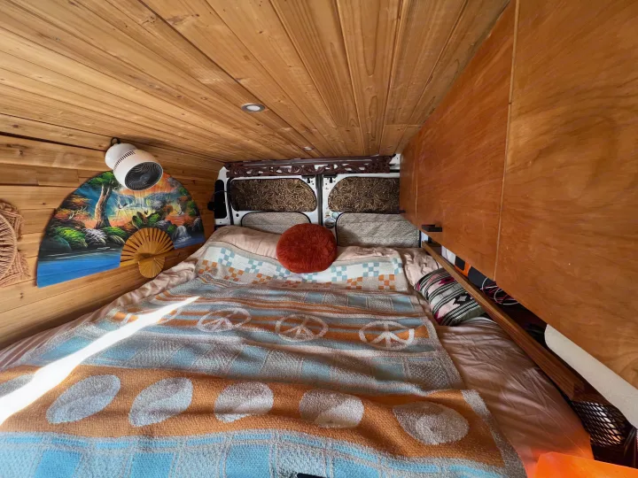 Cozy RV rental interior featuring wooden walls, a comfy bed with a peace-sign blanket, overhead fan, and storage.. Dodge Ram Promaster 2500 2017