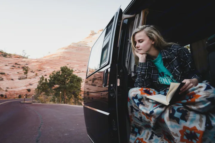 Relaxing RV rental moment: Young woman reading in van door, cozy in a blanket, overlooking scenic desert.. Volkswagen Eurovan MV 2002