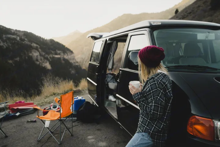 Scenic mountain camping with an RV rental. A woman sips coffee beside her black camper van and outdoor gear.. Volkswagen Eurovan MV 2002