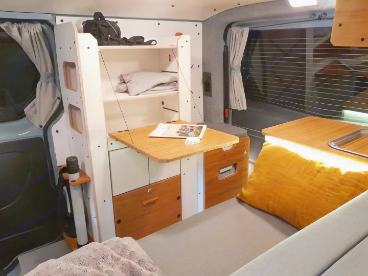 Inside an RV rental, showing a modular camper van interior with a fold-down bamboo table, comfy bed, and compact kitchen.. Renault Trafic aménagé avec kit North West de VAN MANIA 2024