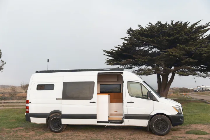 White Sprinter camper van for rv rental, ready for travel, open door reveals custom interior by a tree.. Mercedes-Benz Sprinter 2014