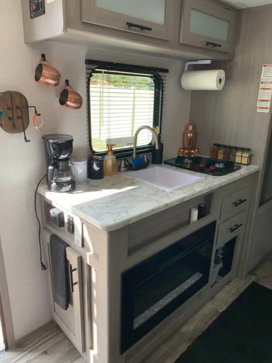 Modern RV rental kitchen featuring light counters, sink, stovetop, coffee maker, and cozy fireplace.. Dutchmen Coleman Lantern LT 2023