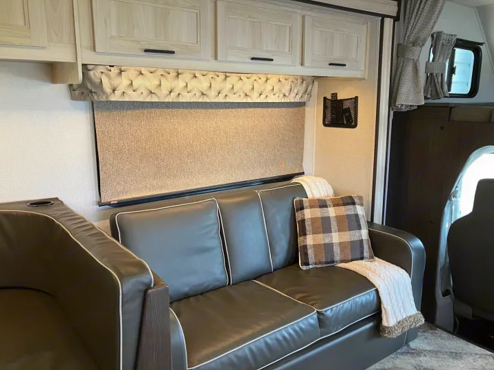 Comfortable RV rental interior: dark couch with white piping, light cabinets, window shade, and remote holder.. Forest River Sunseeker 2020