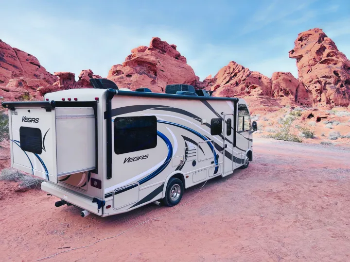 An RV rental, a Vegas motorhome, is parked on red desert earth, dwarfed by majestic red rock formations under a blue sky.. Thor Vegas 2017