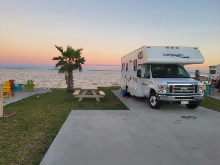 An RV rental is parked at a waterfront site at sunset. A palm tree, picnic table, and colorful chairs overlook the serene water.. Thor Majestic 2017