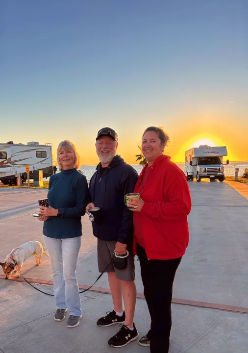Three smiling people with coffee, a dog, and their RVs, enjoying sunrise by the water. Features a Majestic RV rental.. Thor Majestic 2017
