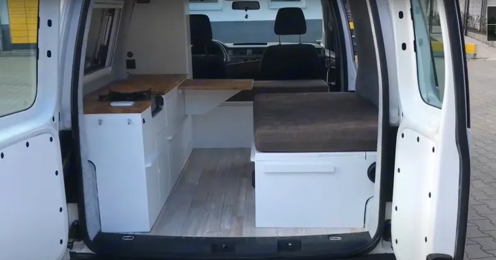 View into an RV rental, showcasing a clean camper van conversion with a compact kitchen, cushioned bed, and wood-look floor.. volkswagen Caddy 2010