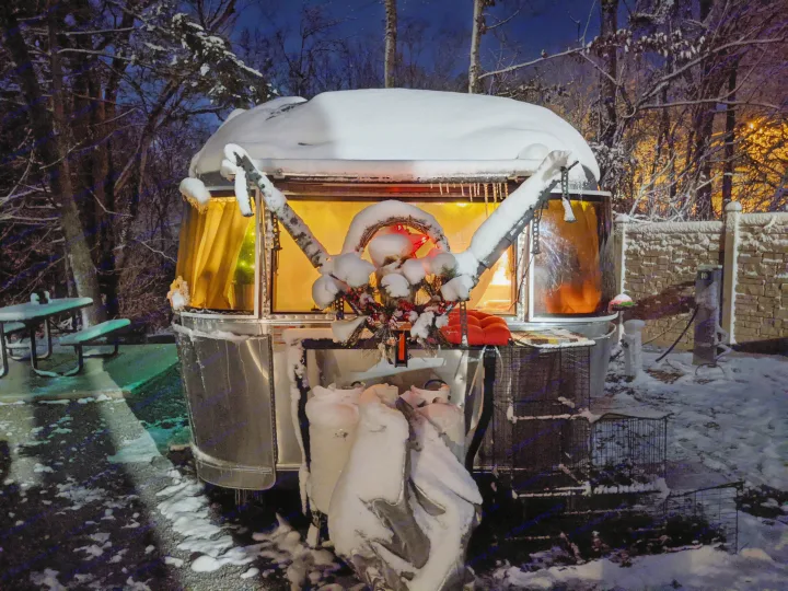 Snow-covered Airstream **rv rental** at night, glowing warmly inside, adorned with holiday decor in a winter landscape.. Airstream International 2015