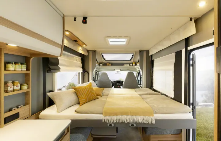 Comfortable RV rental interior: a spacious bed, shelving with jars, and bright light from side windows overlooking nature.. Dethleffs JUST T7052 DBM 2023